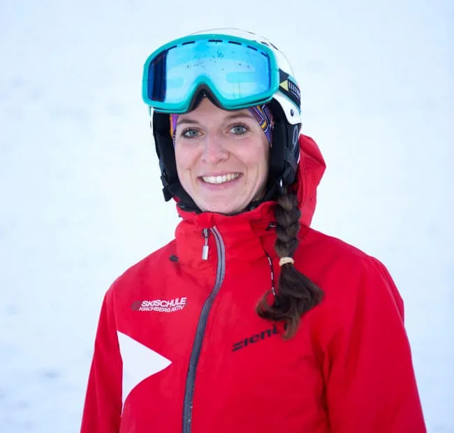 Frau lächelt in Skiausrüstung mit Brille vor verschneitem Hintergrund